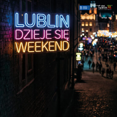 Miasto po zmroku. Na pierwszym planie zbliżenie na kamienicę, na której umieszczony jest podświetlony napis: "Lublin dzieje się weekend". Za kamienicą w tle tłum ludzi idących ulicą i inne kamienice z neonami i światłami okien.