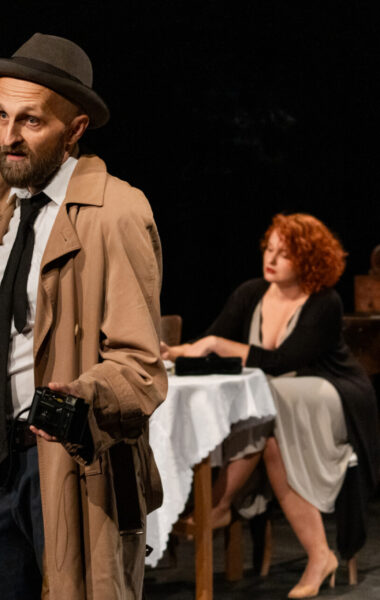 Spektakl teatralny. na scenie, na pierwszym planie mężczyzna w płaszczu, garniturze i kapeluszu. trzyma w ręku pistolet. Za nim na dalszym planie przy stoliku siedzi kobieta w sukience. Dookoła panuje czarny mrok.
