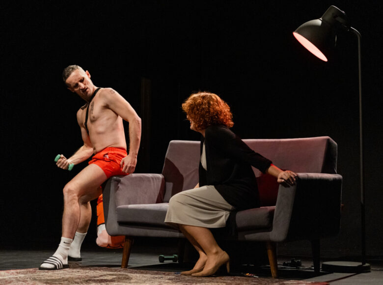 Spektakl teatralny. Na scenie sofa, na sofie siedzi kobieta, a obok niej na oparciu sofy siedzi mężczyzna. Kobieta jest ubrana w sukienkę. mężczyzna ma nago tors, krótkie spodenki gimnastyczne, białe skarpety i sportowe klapki.