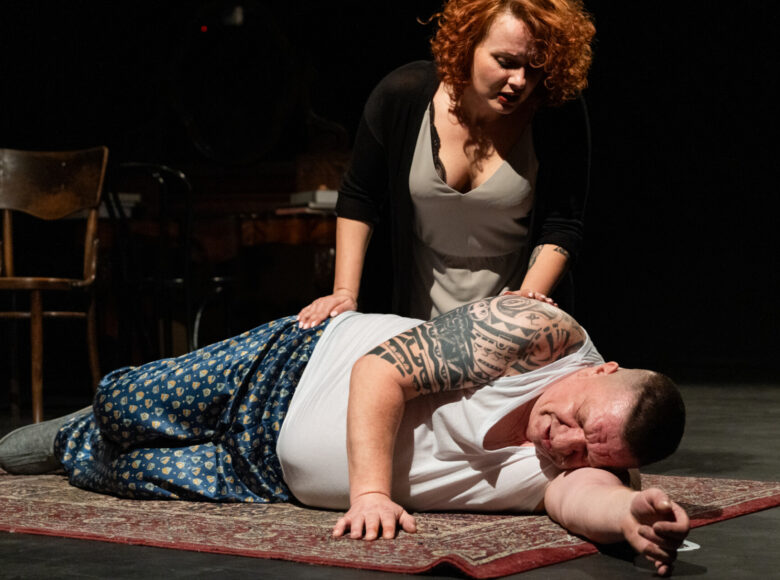Spektakl teatralny. Na podłodze leży stary, gruby mężczyzna w piżamie. nachyla się nad nim klęcząca obok kobieta w sukience. Wokół nich panuje półmrok.