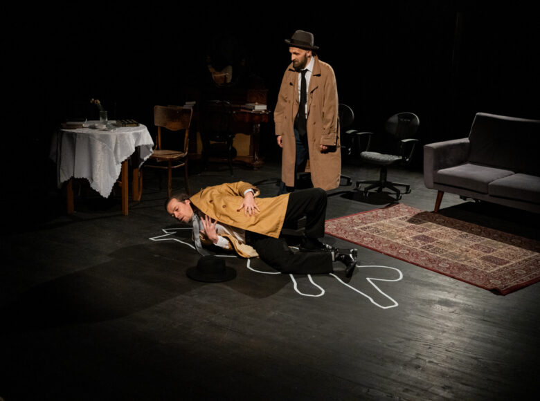 Spektakl teatralny. Na scenie, w półmroku stoi dwóch mężczyzn w płaszczach i garniturach. Jeden leży na podłodze. Wokół niego narysowany jest białą linią obrys sylwetki. Drugi mężczyzna stoi nad pierwszym. Dookoła ustawione są krzesła, stolik i sofa.
