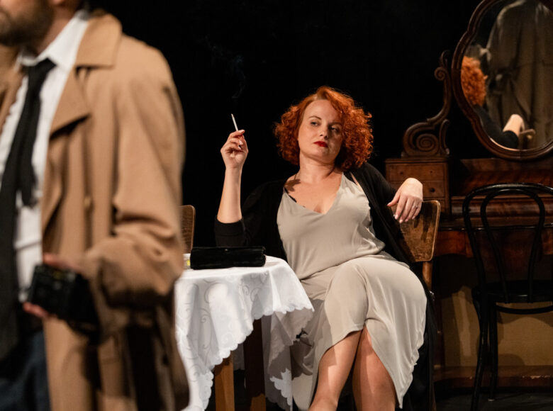 Spektakl teatralny. Na scenie, przy okrągłym stoliku siedzi kobieta z papierosem w dłoni. Patrzy na stojącego obok mężczyznę w płaszczu.
