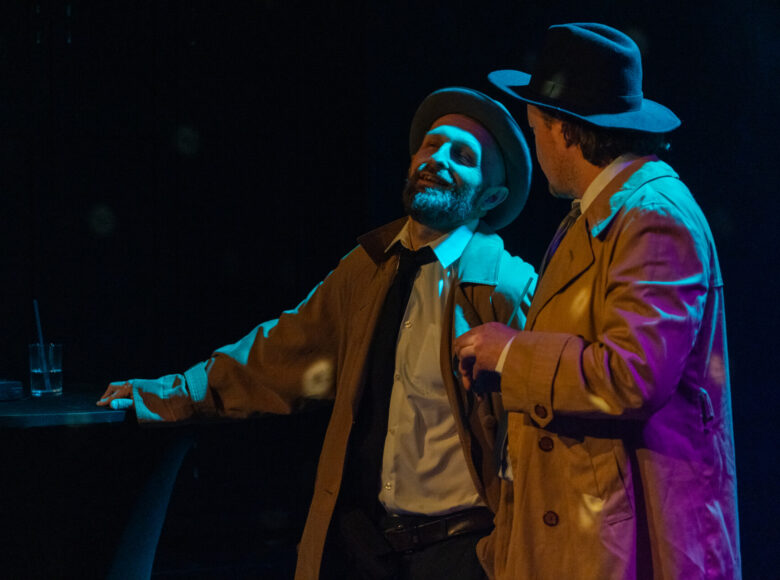 Spektakl teatralny. Na scenie dwóch mężczyzn w płaszczach, stojących obok siebie. Oświetleni są kolorowym, punktowym światłem. Wokół nich panuje czarna ciemność.
