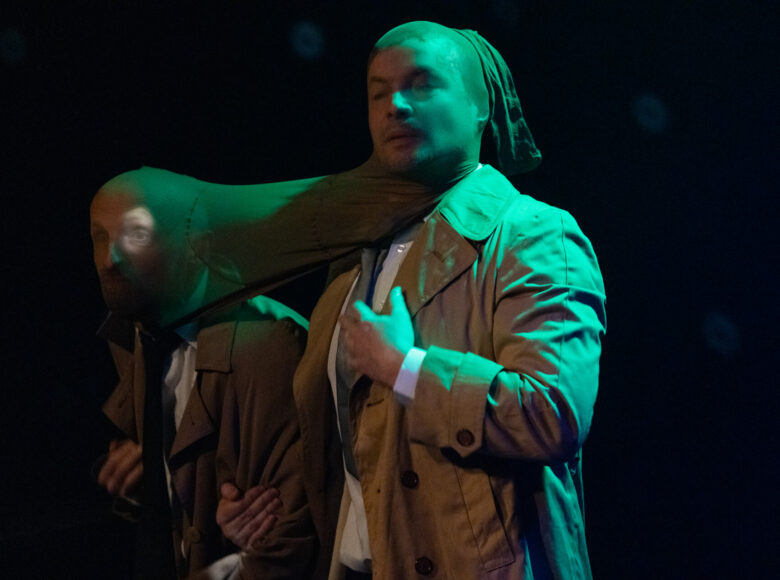 Spektakl teatralny. Zbliżenie na dwóch mężczyzn w płaszczach. Mężczyźni stoją obok siebie. Na głowy i twarze mają naciągniętą jedną nogawkę od cienkich rajstop.