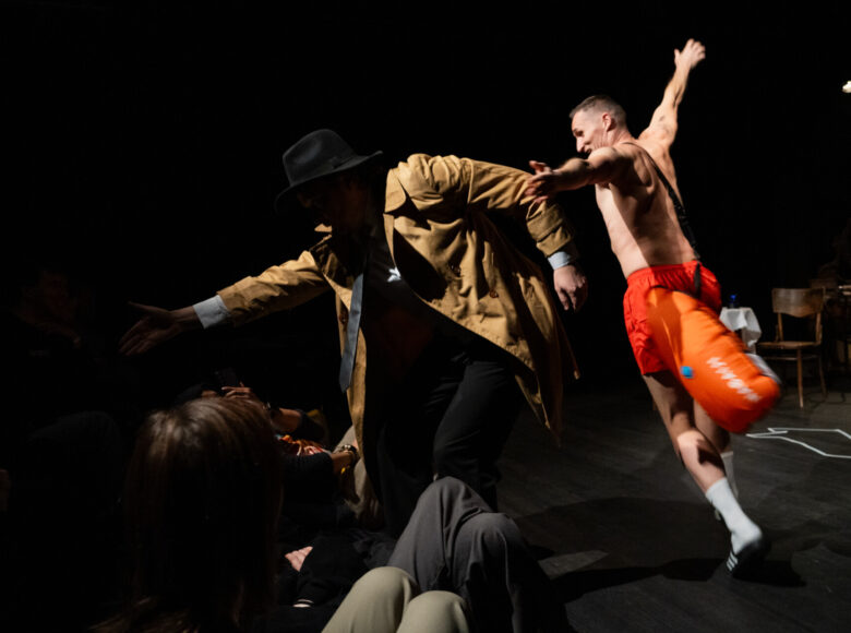 Spektakl teatralny. Na scenie dwóch mężczyzn biegnących w kółko z rozpostartymi rękami. Jeden z nich ubrany jest w płaszcz, garnitur i kapelusz. Drugi ma nagi tors, szorty gimnastyczne, białe skarpety i sportowe klapki.