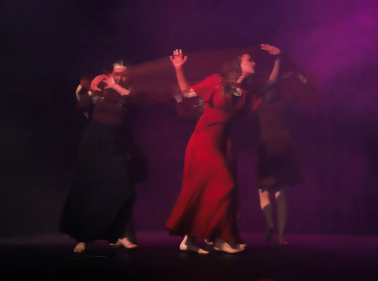 Na ciemnej scenie trzy bose kobiety w rozwianych sukienkach biegają po okręgu, wykonując ekspresyjne gesty rękami.