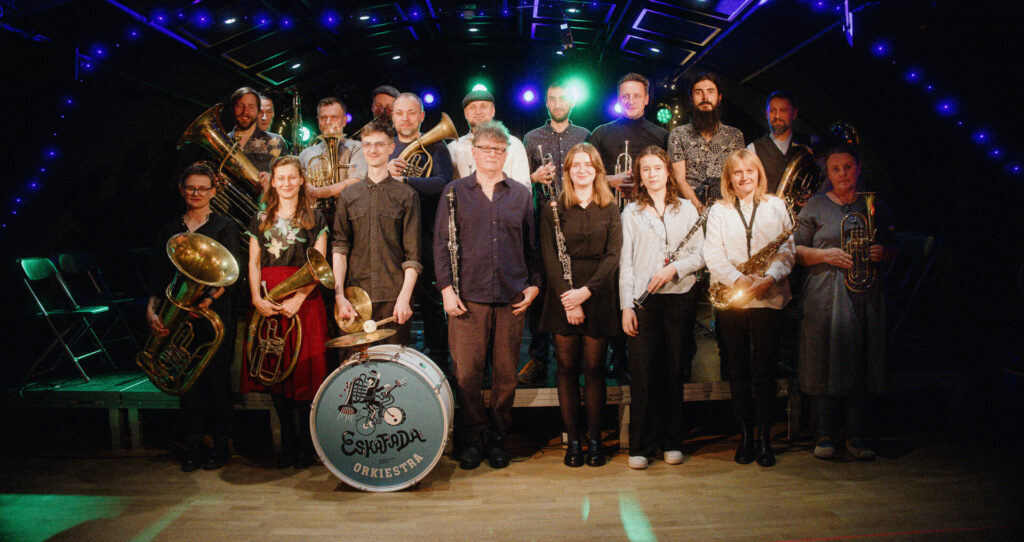 Na scenie grupa kilkunastu osób płci obojga stoi z instrumentami przodem do nas. Za nimi ciemne tło z kilkoma punktami niebieskiego światła.