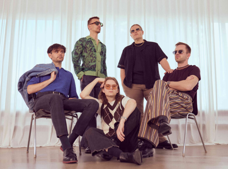 W pomieszczeniu grupa kilku młodych mężczyzn pozuje do fotografii. Ustawienie są na tle okna zasłoniętego półprzezroczystymi zasłonami, przez które wpada światło. Jeden z mężczyzn siedzi na podłodze, dwaj siedzą na krzesłach po jego obu bokach. Za nimi stoi dwóch kolejnych mężczyzn. Mężczyzna siedzący na podłodze patrzy prosto na nas. Pozostali patrzą gdzieś w dal. Wszyscy mają bardzo poważne miny.