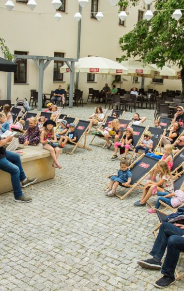Lato. Wirydarz Centrum Kultury w Lublinie. Aktor Dariusz Jeż siedzi na murku i czyta na głos książkę. Przed nim na leżakach siedzą zasłuchani w jego słowa dorośli i dzieci.