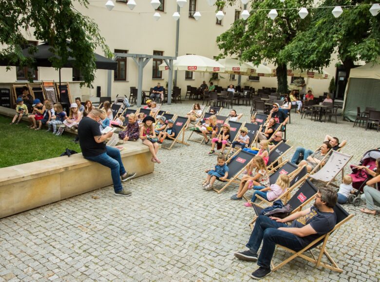 Lato. Wirydarz Centrum Kultury w Lublinie. Aktor Dariusz Jeż siedzi na murku i czyta na głos książkę. Przed nim na leżakach siedzą zasłuchani w jego słowa dorośli i dzieci.