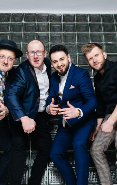 Sześciu elegancko ubranych mężczyzn pozuje do fotografii. Są młodzi i szczupli. Pozują na tle okna w ceglanej ścianie. Okno zrobione jest z luksferów. Mężczyźni sfotografowani są od dołu. Patrzą prosto w obiektyw, pochylając się do przodu.