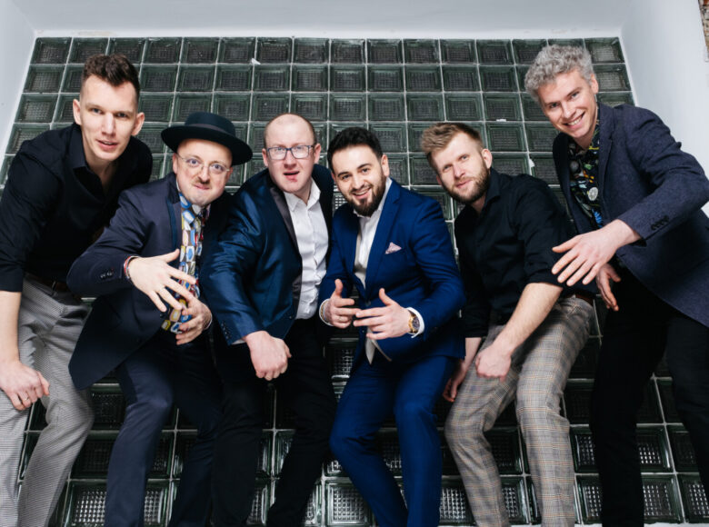 Sześciu elegancko ubranych mężczyzn pozuje do fotografii. Są młodzi i szczupli. Pozują na tle okna w ceglanej ścianie. Okno zrobione jest z luksferów. Mężczyźni sfotografowani są od dołu. Patrzą prosto w obiektyw, pochylając się do przodu.