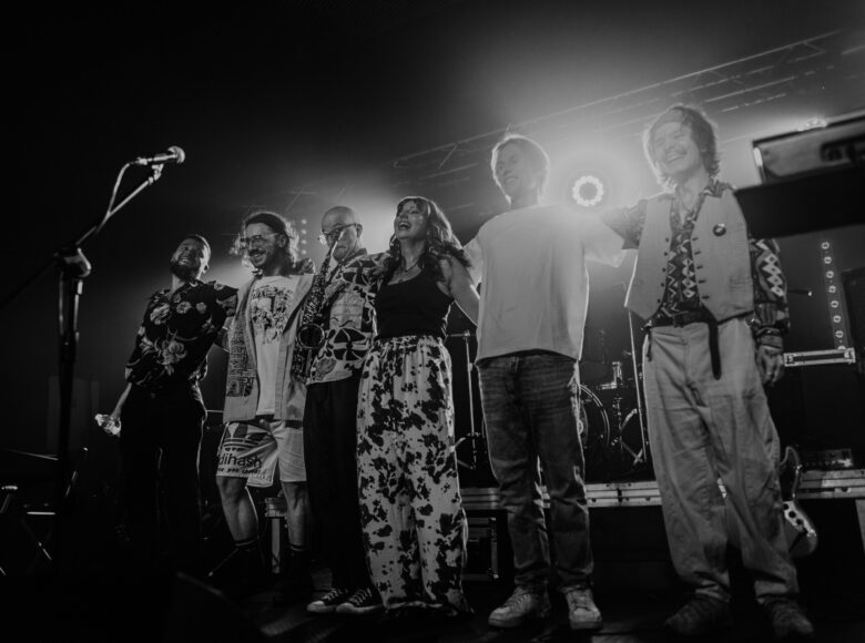 Fotografia czarno biała. Na scenie kobieta i kilku mężczyzn kłaniają się publiczności, stojąc w rzędzie. Jeden z mężczyzn trzyma w ręku saksofon.