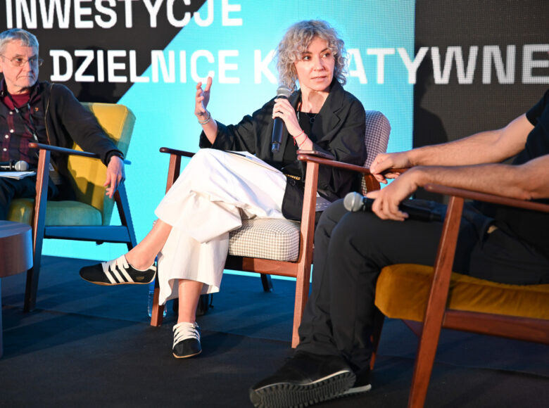 Panel dyskusyjny podczas konferencji. Na scenie na fotelach siedzą obok siebie trzy osoby, kobieta i dwóch mężczyzn. Kobieta, gestykulując, mówi coś do mikrofonu, który trzyma w dłoni.