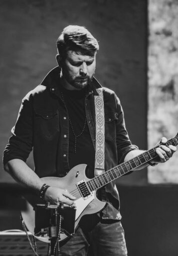 Fotografia czarno biała. Na scenie stoi mężczyzna i gra na gitarze elektrycznej.