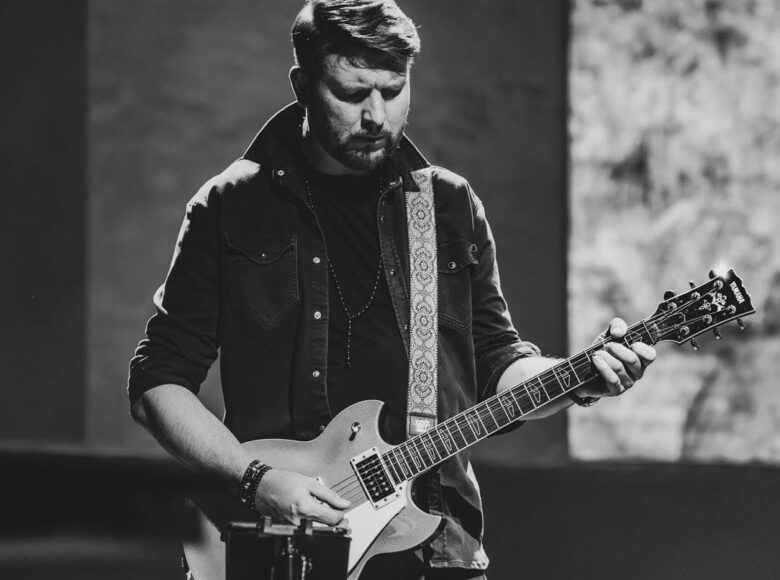 Fotografia czarno biała. Na scenie stoi mężczyzna i gra na gitarze elektrycznej.