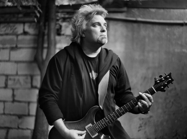 Fotografia czarno białą. Na pierwszym planie stoi mężczyzna grający na gitarze elektrycznej. Patrzy gdzieś w górę, poza kadr. Za jego plecami widzimy mur i drzewo rosnące przy murze.