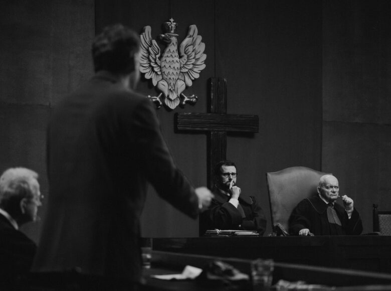 Sala rozpraw sądowych. Na pierwszym planie stoi zeznający mężczyzna. W tle siedzą dwaj mężczyźni w togach.
