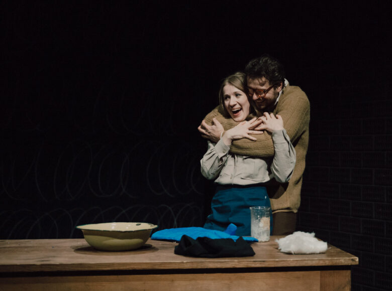 Przedstawienie teatralne. Na scenie przy stole stoją kobieta i mężczyzna, obejmując się.