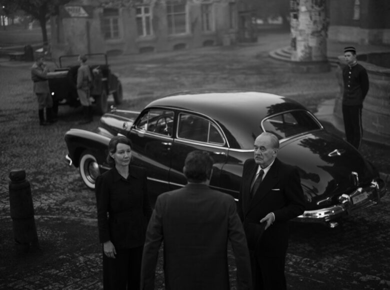 Kadr z filmu czarno biały. Na miejskim placu stoi samochód z połowy dwudziestego wieku. Obok niego kilku mężczyzn i kobieta w ubraniach także z połowy dwudziestego wieku. W tle samochód wojskowy i dwóch żołnierzy.