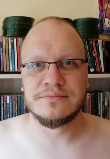 Zbliżenie na twarz i nagie ramiona młodego mężczyzny. Mężczyzna ma brodę i okulary. Patrzy prosto na nas. Za nim widzimy półkę z książkami.