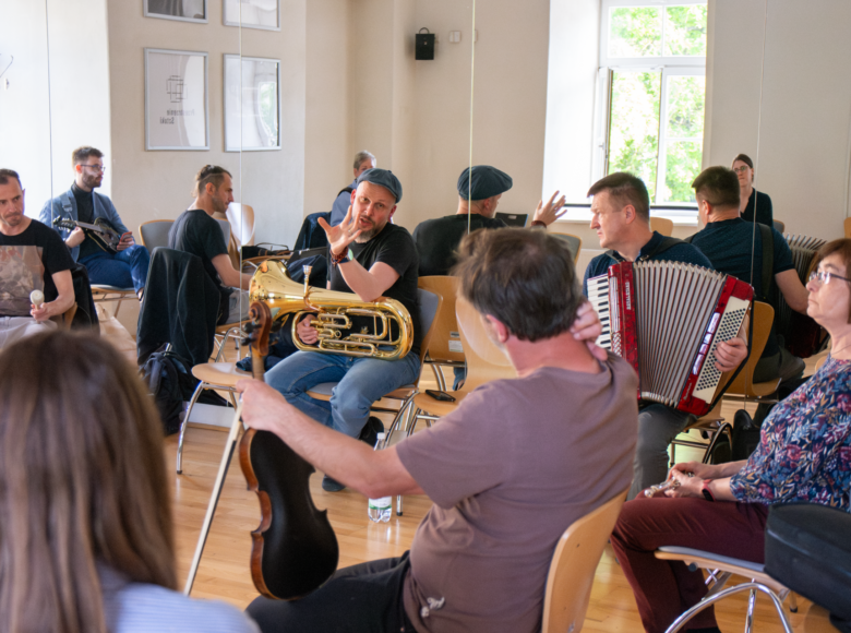 Kilka osób gra na różnych instrumentach muzycznych, siedząc w sali na krzesłach.