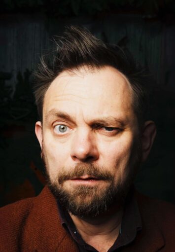 Fotograficzny portret mężczyzny w średnim wieku, z wąsami i brodą. Mężczyzna patrzy prosto na nas, przymykając jeno oko.