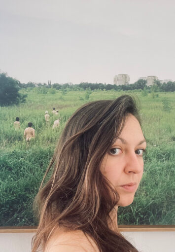Zbliżenie na twarz młodej kobiety. Kobieta stoi na tle ściany, na której wisi fotografia przedstawiająca grupę nagich ludzi spacerujących w zaroślach, na tle osiedla mieszkaniowego.