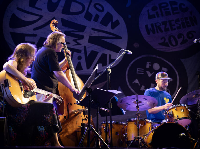Na scenie muzyczka z gitarą i dwóch muzyków, jeden z kontrabasem, a drugi z perkusją.