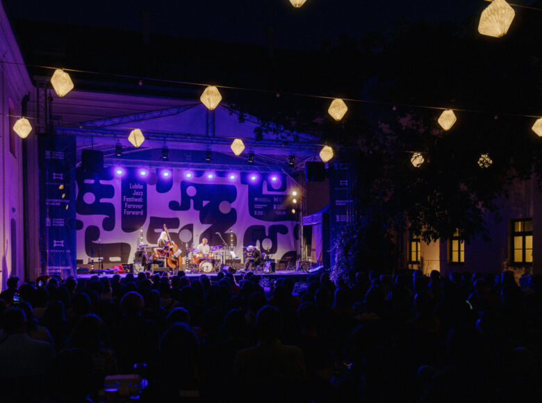 Nocny koncert na wirydarzu Centrum Kultury w Lublinie. Na pierwszym planie widzimy od tyłu widownię, a w głębi scenę i na niej muzyków.